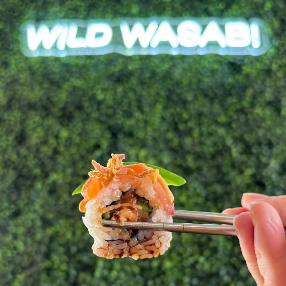 Close-up of a sushi roll held with chopsticks in front of a green wall with 'Wild Wasabi' sign.