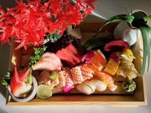 A wooden tray holds an assortment of sashimi and sushi garnished with lemon, wasabi, and greens. A small vase with leaves and red decorative foliage is also placed on the tray.