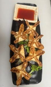 Five star-shaped pastries arranged on a black rectangular plate with a small dish of red sauce at the top, garnished with lettuce leaves.