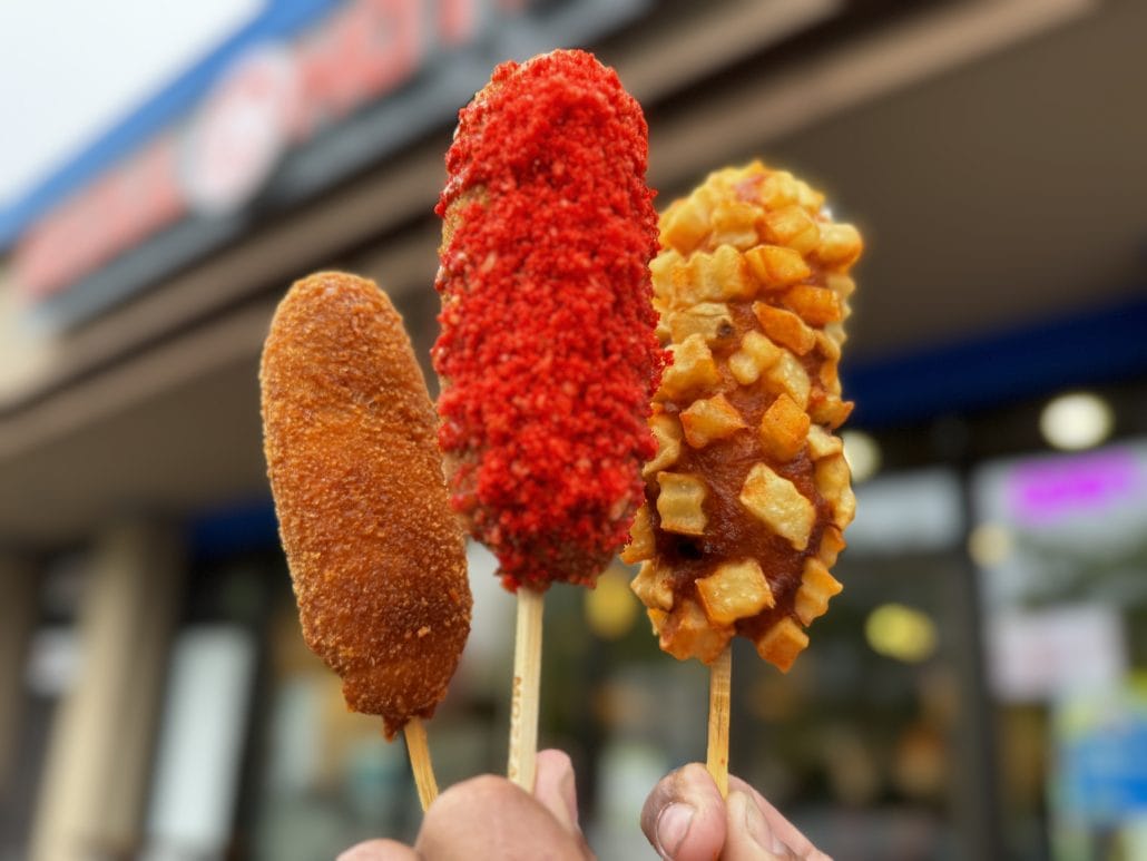 Three colorful corn dogs on sticks held up in front of a food stand in Lynnwood, WA.